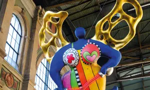 Silhouette d'une Nana colorée de Niki de Saint Phalle, avec des ailes dorées, suspendue dans un grand hall.