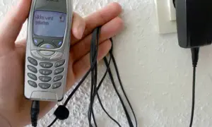 Dłoń trzyma starą Nokię, która ładuje się przez kabel. Na ekranie telefonu widnieje napis "Akku wird geladen". To klasyczny przykład technologii 3G z dawnych lat.