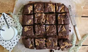 Pyszne brownie z masłem orzechowym, pokrojone w kostkę, czeka na degustację. Obok szklanka mleka i delikatne białe kwiaty.