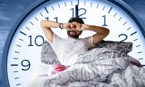 Man in bed rubs his eyes, looking tired. A large clock face behind him shows it's just past 6 AM, suggesting only six hours of sleep.