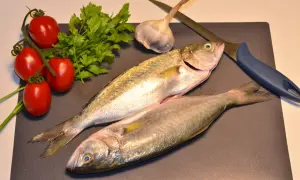 Due pesci serra freschi, pomodorini, prezzemolo, aglio e un coltello su un tagliere, pronti per una cena deliziosa.