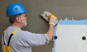 Pracownik w niebieskim kasku nakłada zaprawę z siatką na styropian. Poznaj cenę za położenie siatki z klejem.