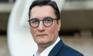 Olivier Dartigolles, un homme aux lunettes bleues, pose pour un portrait. Il est vêtu d'un costume sombre, évoquant une image de famille et de professionnalisme.