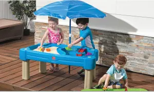 Drei Kinder genießen Sommerspiele im Sand- und Wasserspieltisch unter einem blauen Sonnenschirm.