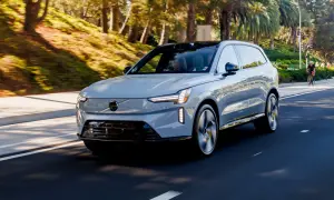 Szary Volvo EX90 jedzie po słonecznej drodze, otoczony zielenią i palmami.
