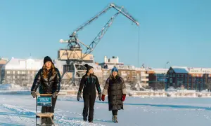 Tre kvinnor njuter av en vinterdag och utforskar Luleå på isen, med en stor kran och snötäckta byggnader i bakgrunden.