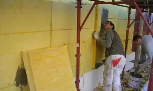 Pracownicy montują izolację na ścianie. Jeden z nich sprawdza pion za pomocą poziomicy, dbając o idealny wzór na lambdę.