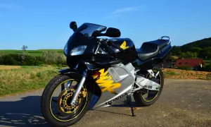Czarny motocykl Honda NSR z żółtymi akcentami na tle zielonych pól i błękitnego nieba.