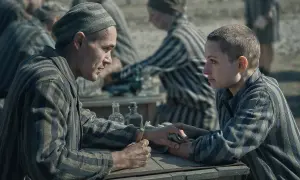Två fångar i randiga uniformer möts vid ett bord. Mannen håller en nål, redo att tatuera kvinnan. Scen från filmen "Tatueraren i Auschwitz".