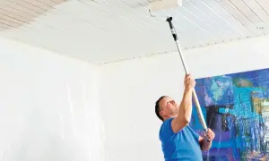 Man målar taket inomhus med en lång roller. En del av taket är redan vitt, medan den andra delen fortfarande är träfärgad. Väggarna är täckta med plast för att skydda dem.