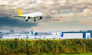Ein Linienflugzeug mit gelbem Leitwerk nähert sich dem Flughafen. Die Linienflug Geschwindigkeit ist beeindruckend, während es über das Schilf und die Gebäude fliegt.