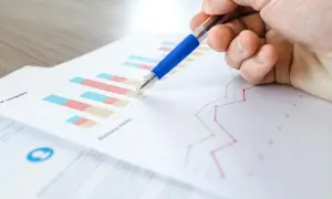 Dłoń z niebieskim długopisem wskazuje na wykres słupkowy i liniowy, prawdopodobnie wygenerowane w **ms excel**. Analiza danych biznesowych.