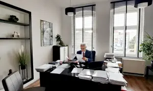 Mężczyzna w garniturze siedzi przy biurku w gabinecie, przeglądając dokumenty.