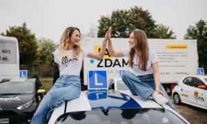 Dwie uśmiechnięte dziewczyny świętują zdanie egzaminu na prawo jazdy kat. B, siedząc na dachu samochodu szkoleniowego.