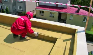 Pracownik w czerwonym kombinezonie wykonuje ocieplenie wykuszu od zewnątrz, układając płyty izolacyjne.