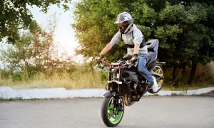 Motocyklista w kasku wykonuje jazdę na tylnym kole na asfalcie, otoczony zielenią.