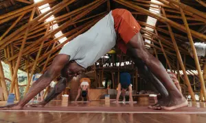 Un homme en posture de chien tête en bas, cherchant à redresser son dos. D'autres pratiquent le yoga dans un studio en bambou.