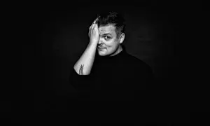 Czarno-białe zdjęcie. Mężczyzna z tatuażem na ramieniu, w czarnym golfie, zasłaniający dłonią jedno oko. Wygląda jak król piosenkarz.