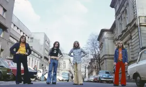 Czterech mężczyzn w stylu lat 70. na ulicy. Jeden z nich ma na koszulce napis "nie wierz nigdy kobiecie tekst".