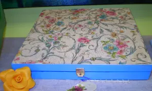 Caja azul con tapa decorada con papel decoupage floral. Un detalle de rosa amarilla y etiqueta floral completan la escena.