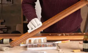 Persona con guantes blancos restaura un mueble de madera, usando un bloque para lijar y kits de retoque.