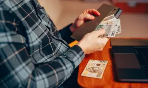 Osoba w kratę liczy pieniądze, przygotowując donos do urzędu skarbowego. Obok leży laptop.