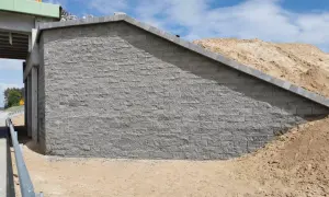 Mur oporowy z szarej kostki brukowej, obok nasypu ziemnego i fragmentu wiaduktu.