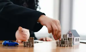 Dłoń układa monety obok skarbonki, modelu domu i samochodu.