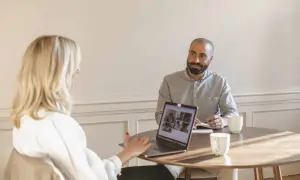 Två personer vid ett runt bord, en kvinna med blont hår och en man med skägg. De har en laptop och anteckningsblock, vilket tyder på ett konstruktivt samtal.