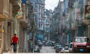 Photographie de rue d'une rue cubaine animée, avec des bâtiments colorés et délabrés, des voitures anciennes et un homme marchant seul.