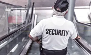 Pracownik ochrony w białej koszulce z napisem "SECURITY" stoi na ruchomych schodach, czuwając nad bezpieczeństwem.