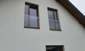 Nowoczesny portfenetr z drewnianymi ramami i szklaną balustradą przy oknach na białej elewacji.