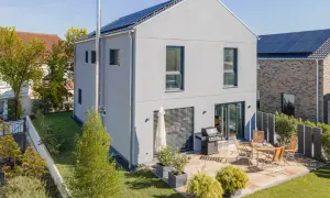 Ein modernes, schmales Haus mit Solaranlage auf dem Dach und einer Terrasse mit Grill und Sitzmöbeln.