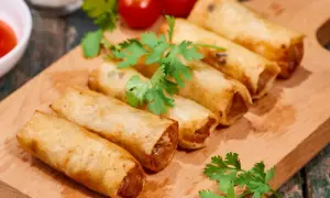 Chrupiące spring rolls na desce, ozdobione kolendrą i pomidorkami. Idealne na przekąskę.