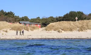 Drei Angler spazieren am Strand von Bornholm entlang, vorbei an roten Häusern und Dünen.