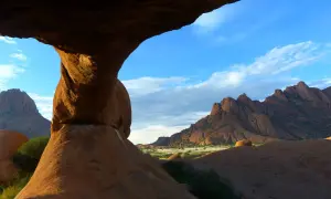 Felsformationen der Spitzkoppe in Namibia, mit einem Felsentor im Vordergrund und zerklüfteten Bergen am Horizont unter blauem Himmel.