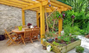 Un pergolato terrazzo fai da te accoglie un tavolo in legno e sedie, circondato da aiuole fiorite e vegetazione lussureggiante.