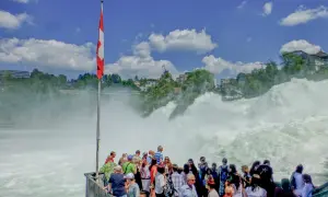 Menschen bestaunen den tosenden Rheinfall. Eine Schweizer Fahne weht im Wind. Der **Rheinfall Eintritt** ist ein beeindruckendes Naturschauspiel.
