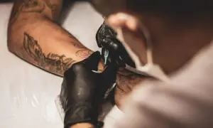Artysta w czarnej rękawiczce tworzy tatuaż na barku klienta. Widać kontury nowego wzoru i fragmenty już istniejących tatuaży.