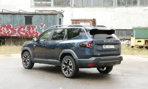 Ciemnoniebieska Dacia Bigster, model hybrydowy, zaparkowana na tle opuszczonej fabryki z wagonami kolejowymi pokrytymi graffiti.