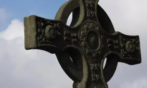 Kamienny krzyż celtycki z misternymi zdobieniami na tle błękitnego nieba z chmurami.