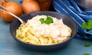 En skål med tagliatelle toppad med krämig ost och skinksås, garnerad med persilja. Två gula lökar syns i bakgrunden.