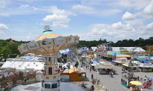 Buntes Treiben auf dem Volksfest Ingolstadt mit Kettenkarussell, Fahrgeschäften und Buden unter blauem Himmel mit Wolken.