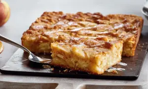 Saftige schwedische Apfeltorte, mit Äpfeln belegt und mit Puderzucker bestäubt, auf einem dunklen Brett mit einer Gabel.