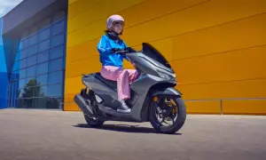 Kobieta w różowych spodniach i niebieskiej kurtce jedzie na szarym skuterze Honda PCX 125. Wady tego modelu mogą dotyczyć np. zawieszenia.