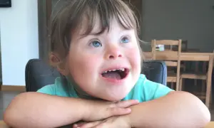 Una bambina down con occhi azzurri e un sorriso radioso, appoggiata al tavolo con le braccia incrociate.