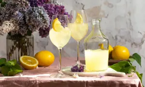 Zwei Gläser Limoncello Spritz mit Zitrone, eine Flasche Limoncello, Zitronen und Flieder auf rosafarbenem Tischtuch.
