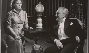 Para z dramatu Ibsena, ona w eleganckiej sukni, on w smokingu, siedzą naprzeciw siebie.