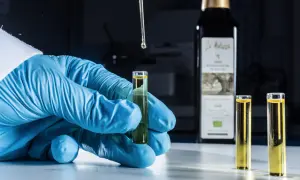 Analisi in laboratorio per verificare i perossidi nell'olio extravergine di oliva. Goccia di olio cade da pipetta in provetta tenuta da mano con guanto blu.