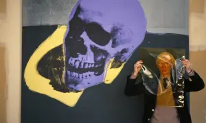 Andy Warhol trzyma przezroczystą folię przed fioletowo-żółtym obrazem czaszki.